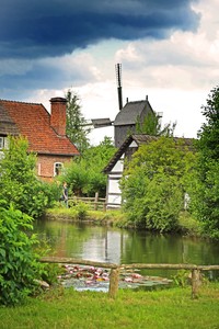 Vorne ist ein Teich. Dahinter steht eine Wind-Mühle. Neben der Wind-Mühle steht ein Fachwerk-Haus.