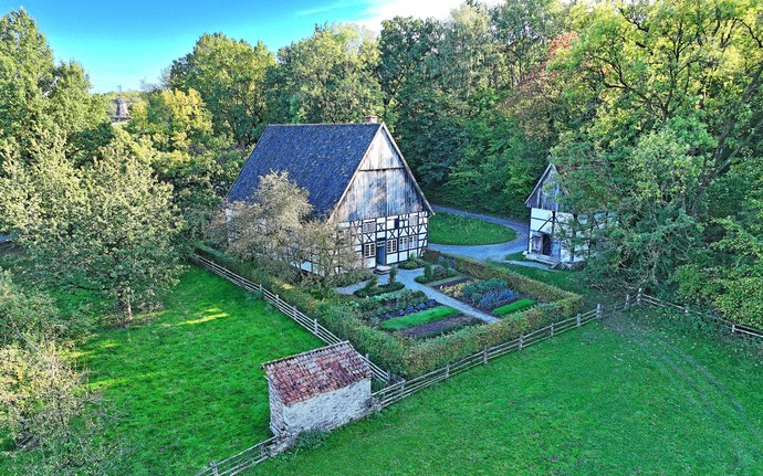 Ein Fachwerkhaus vor Bäumen, davor ein Gemüsegarten und eine Obstbaumwiese.