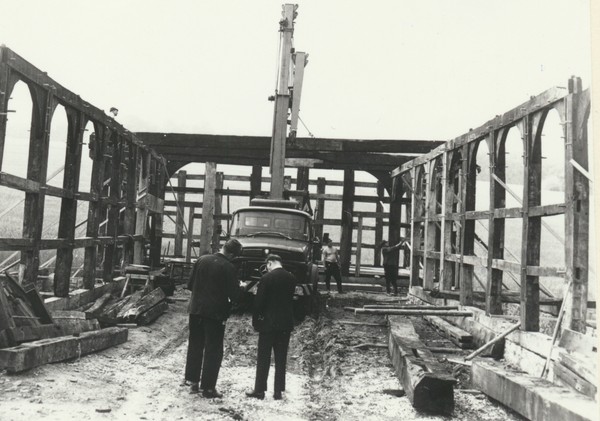 Schwarzweiß-Bild eines Fachwerkgerippes, in der Mitte ein LKW aus den 1960er Jahren mit Kran, davor zwei Arbeiter.