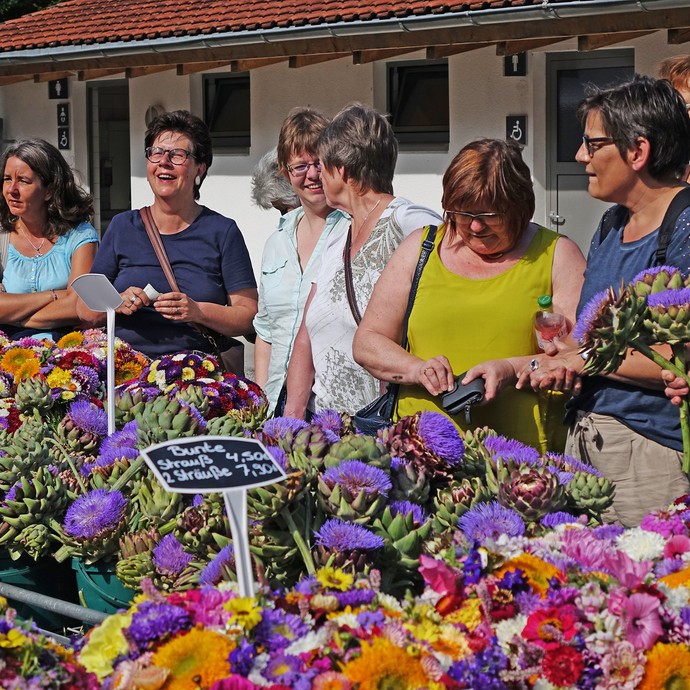 An einem Stand mit vielen bunten Blumensträußen stehen sechs Frauen, lachen, unterhalten sich und kaufen Sträuße. (vergrößerte Bildansicht wird geöffnet)