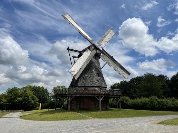 eine Windmühle mit bespannten Flügeln und umlaufender Galerie vor blauem Himmel