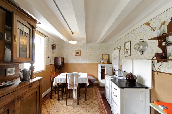 Eine Küche, eingerichtet wie in den 19509er Jahren: links ein Holzschrank, in der Mitte ein Tisch mit Stühen, rechts ein alter Herd und ein Kühlschrank.