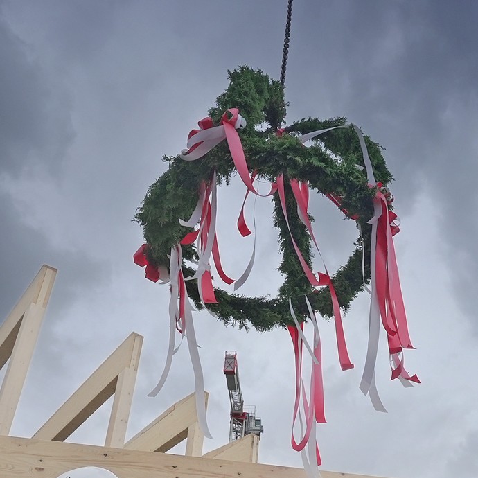 Ein Richtkranz mit roten und weißen Bändern hängt über einem Dachstuhl aus Holz. (vergrößerte Bildansicht wird geöffnet)