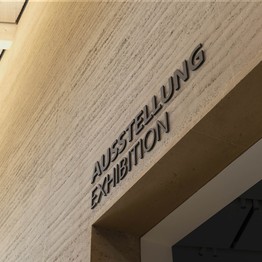 Die Worte "Ausstellung" und "Exhibition" stehen an einer Lehmwand.