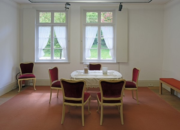 vier rote Stühle stehen um einen Tisch, im Hintergrund zwei Fenster mit weißen Gardinen