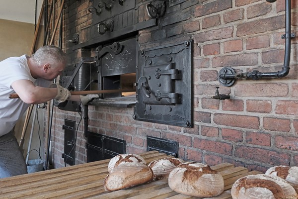 Ein Bäcker steht vor einem geöffneten alten Ofen aus Backstein, im Vordergrund liegen gebackene Brotlaibe auf einem Holzbrett