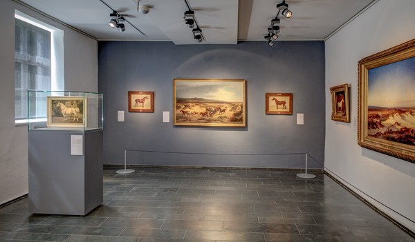 Ein Ausstellungsraum, in dem mehrere Gemälde hängen. Links ein kleineres Gemälde in einer Glasvitrine.