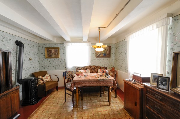 Wohnzimmer mit Einrichtung der 1950er Jahre: In der Mitte ein Tisch mit Stühlen und Sofa, links ein Schrank und ein Holzofen, rechts ein Fernseher auf einer Anrichte.